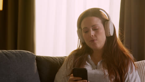 Woman-Wearing-Wireless-Headphones-Sitting-On-Sofa-At-Home-Streaming-Music-From-Mobile-Phone-3